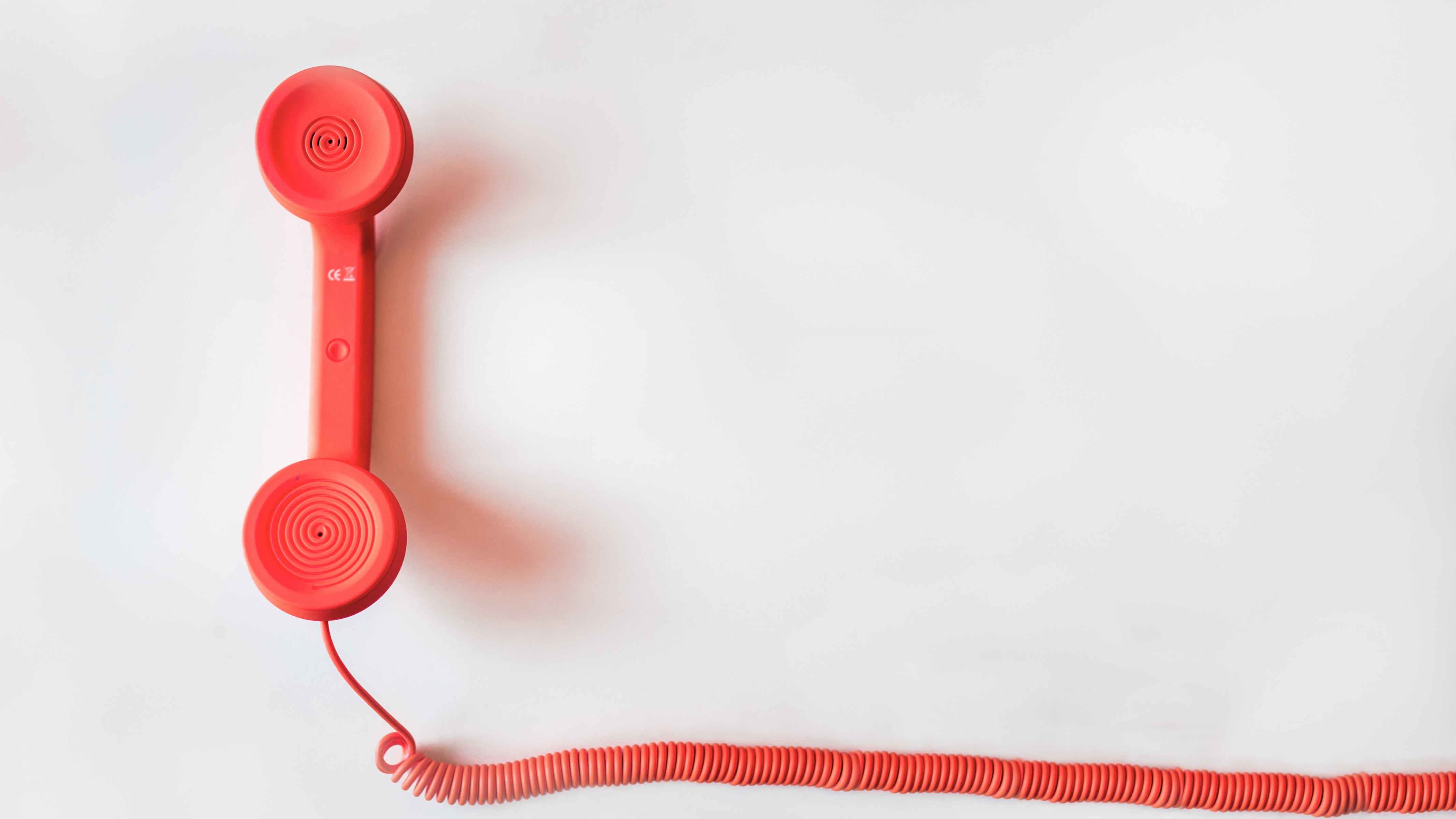 A bright red telephone handset resting on a light grey studio surface, its coiled cord trailing across the frame